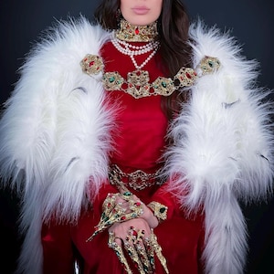 Puede incluir: Un conjunto regio con un vestido rojo, una estola de piel sintética blanca y joyas de oro ornamentadas. Las joyas incluyen un collar, un collar, un cinturón, pulseras y cubiertas de uñas elaboradas, todas adornadas con piedras preciosas rojas y verdes. Una corona completa el look.