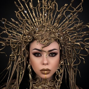 May include: A woman wearing a gold snake crown and necklace. The crown is made of many small snakes that are intertwined. The necklace is also made of snakes, but they are arranged in a more circular pattern. The woman has dark hair and dark eyes. She is wearing gold makeup.
