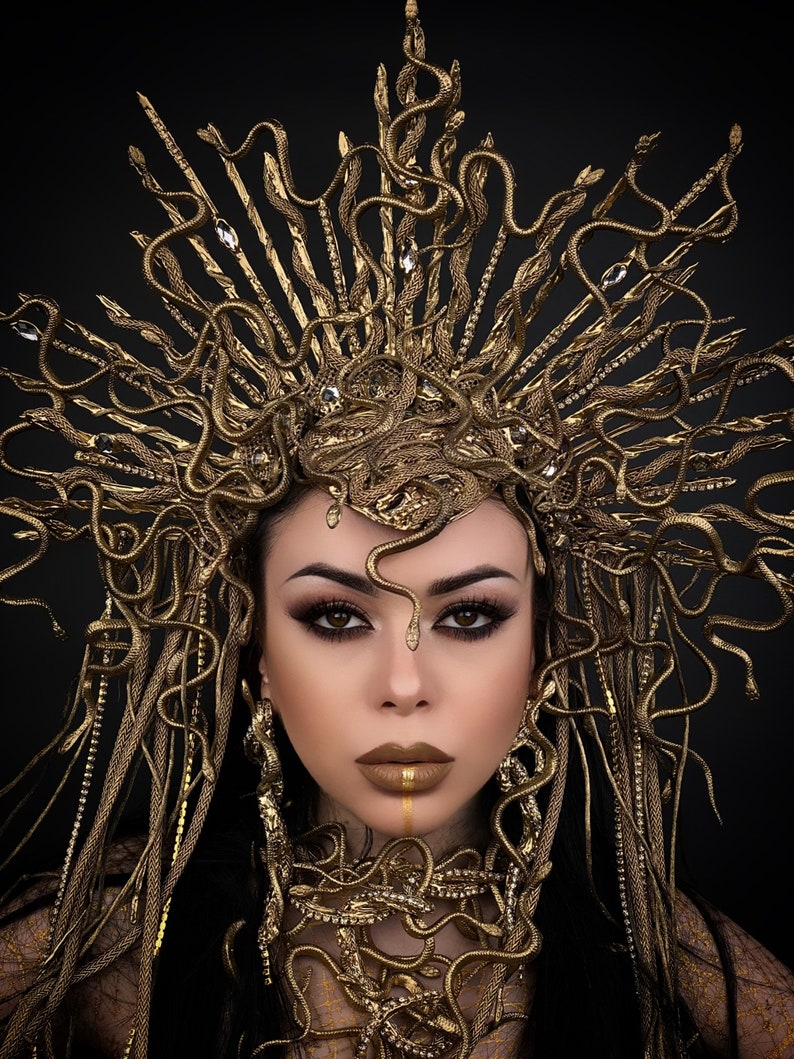 May include: A woman wearing a gold snake crown and necklace. The crown is made of many small snakes that are intertwined. The necklace is also made of snakes, but they are arranged in a more circular pattern. The woman has dark hair and dark eyes. She is wearing gold makeup.