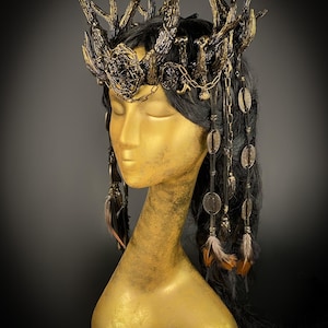 May include: A gold mannequin head showcases a dark-haired crown with antler-like structures, chains, and dangling embellishments. The crown features a central ornate design and is adorned with feathers and circular pendants. The overall aesthetic is fantasy-inspired.