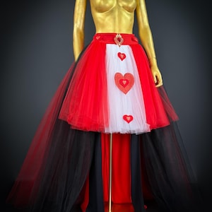 May include: A red, white, and black costume skirt with heart accents. The skirt features layers of red and white tulle, with black tulle cascading from the back. A gold mannequin displays the skirt, which has a red waistband with a heart-shaped embellishment.