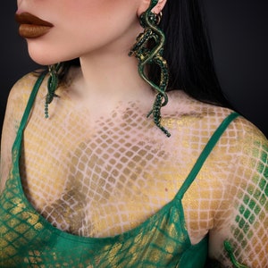 May include: A woman wearing a green top with a snake scale pattern. She is wearing large green snake earrings.