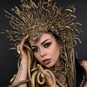 May include: A woman wearing a gold snake crown and a gold snake necklace. The snakes are intertwined around her neck and arms. The woman has dark hair and is wearing gold makeup. The background is black.