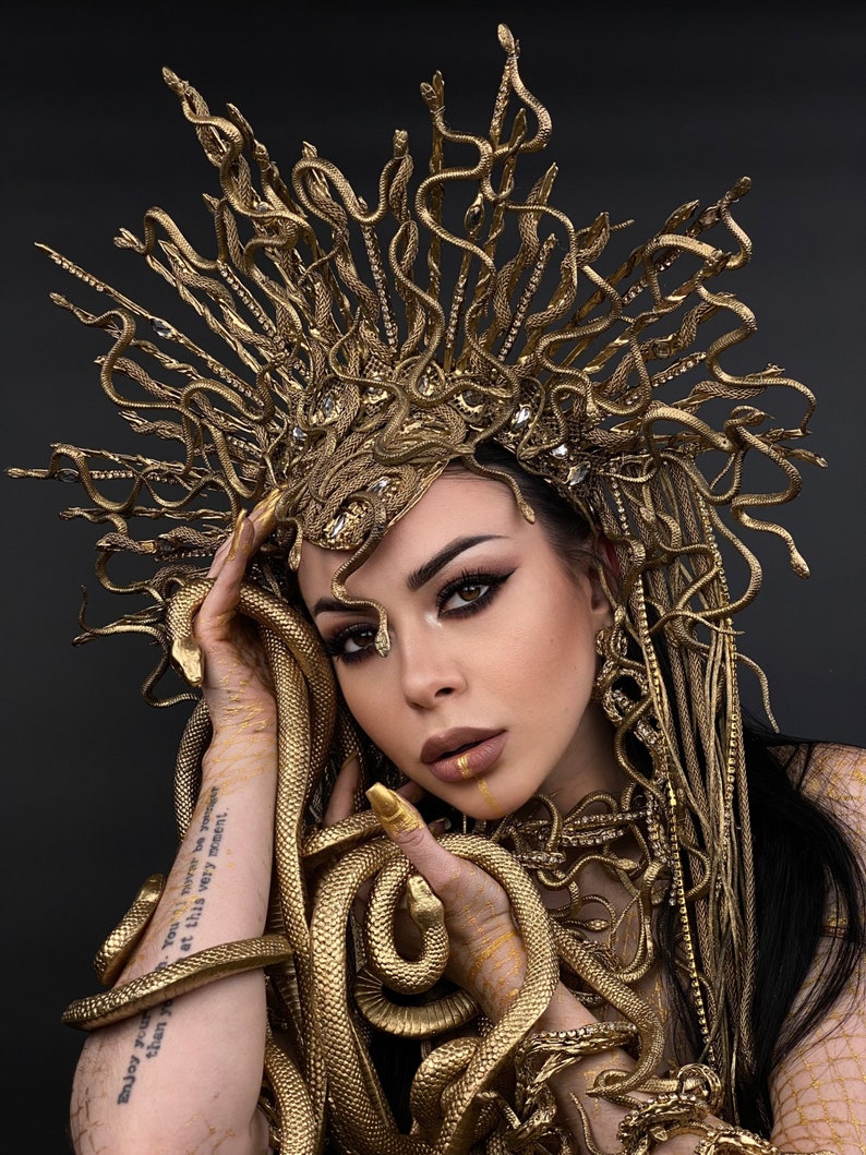 May include: A woman wearing a gold snake crown and a gold snake necklace. The snakes are intertwined around her neck and arms. The woman has dark hair and is wearing gold makeup. The background is black.