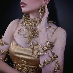 May include: A woman wearing a gold dress and jewelry made of snakes. The jewelry is on her arms, neck, and ears.