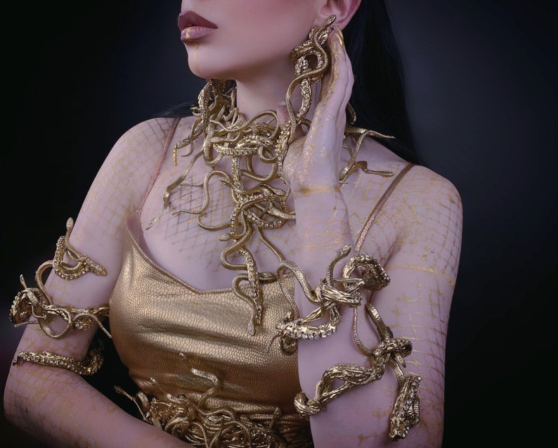 May include: A woman wearing a gold dress and jewelry made of snakes. The jewelry is on her arms, neck, and ears.