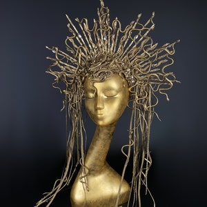 May include: A gold-colored headpiece with a crown of snakes and long, flowing strands of snakes. The headpiece is on a mannequin head.