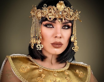 May include: A gold headpiece featuring intricate snake motifs, an ankh symbol, and dangling beaded chains, paired with a wide gold collar necklace adorned with embossed scarab beetles, winged figures, and cobra designs over a white garment.