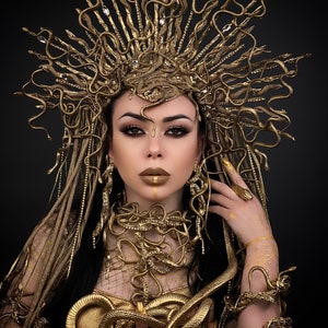 May include: A woman wearing a gold snake crown, necklace, and bracelets. The snakes are intertwined and form a decorative pattern. The woman has dark hair and is wearing gold makeup.