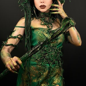 May include: A woman in a green and gold costume with a snake-like staff. The costume is made of fabric and has a snake-like pattern. The staff is made of wood and has a snake head on the top.