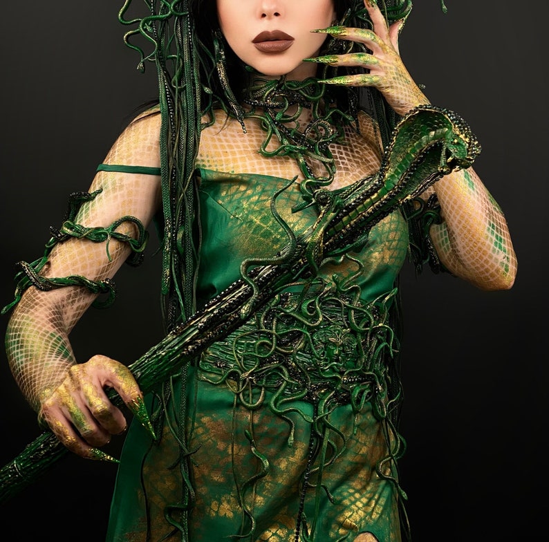 May include: A woman in a green and gold costume with a snake-like staff. The costume is made of fabric and has a snake-like pattern. The staff is made of wood and has a snake head on the top.
