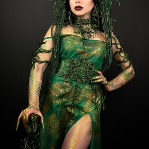 May include: A woman wearing a green and gold dress with a snake-like pattern. The dress has a high neckline and a fitted bodice. The woman is wearing a snake-like headpiece and has green makeup on her eyes and lips.