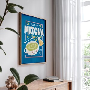 May include: A framed poster with a blue background and the text "JAPAN'S MATCHA" in yellow and white letters. A cartoon illustration of a cup of green tea with a smiling face and a whisk is in the center of the poster. The text "抹茶" is in the lower right corner of the poster.