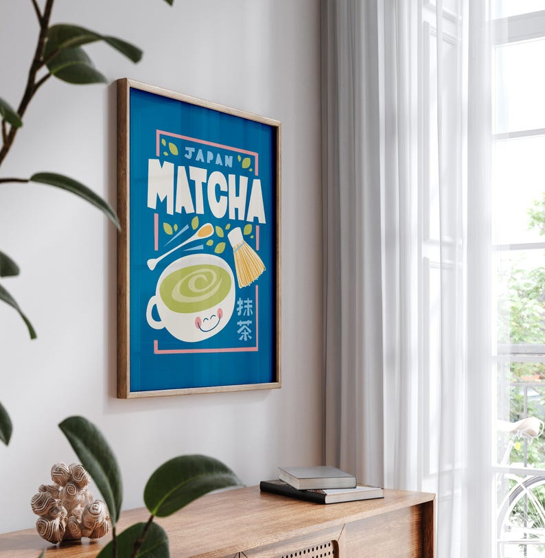 May include: A framed poster with a blue background and the text "JAPAN'S MATCHA" in yellow and white letters. A cartoon illustration of a cup of green tea with a smiling face and a whisk is in the center of the poster. The text "抹茶" is in the lower right corner of the poster.