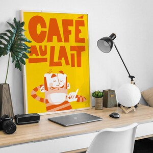 A playful "Café au Lait" poster from Almanac Co Prints sits on a desk with bold yellow and orange colors, featuring a happy character holding a cup of coffee. Framed on a modern desk next to laptop.