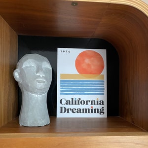 Almanac Co Prints framed poster titled 'California Dreaming' with a red sun, blue waves, and minimalist 1970s design, perfect for home decor. Framed leaning on wooden shelf next to ceramic ornament.