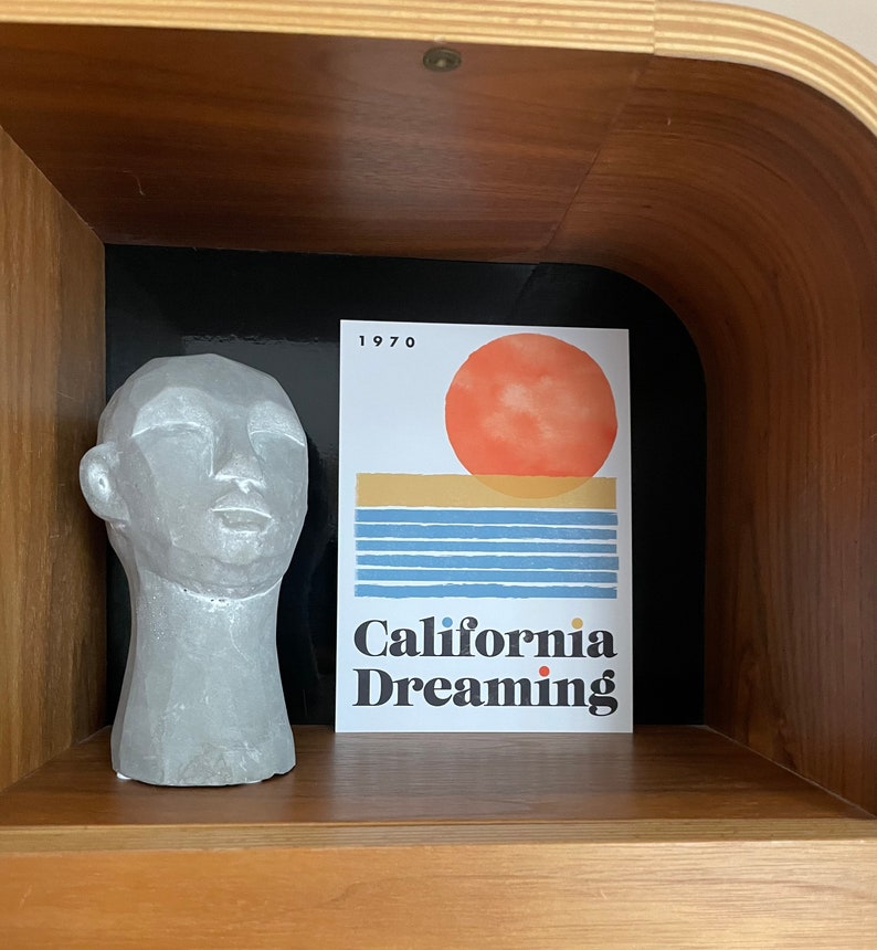 Almanac Co Prints framed poster titled 'California Dreaming' with a red sun, blue waves, and minimalist 1970s design, perfect for home decor. Framed leaning on wooden shelf next to ceramic ornament.