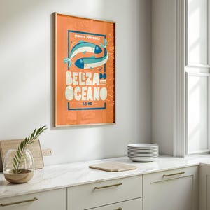 Almanac Co Prints orange poster featuring two colorful fish with the text 'Beleza do Oceano' in bold lettering, Portuguese style. Framed in modern white kitchen.