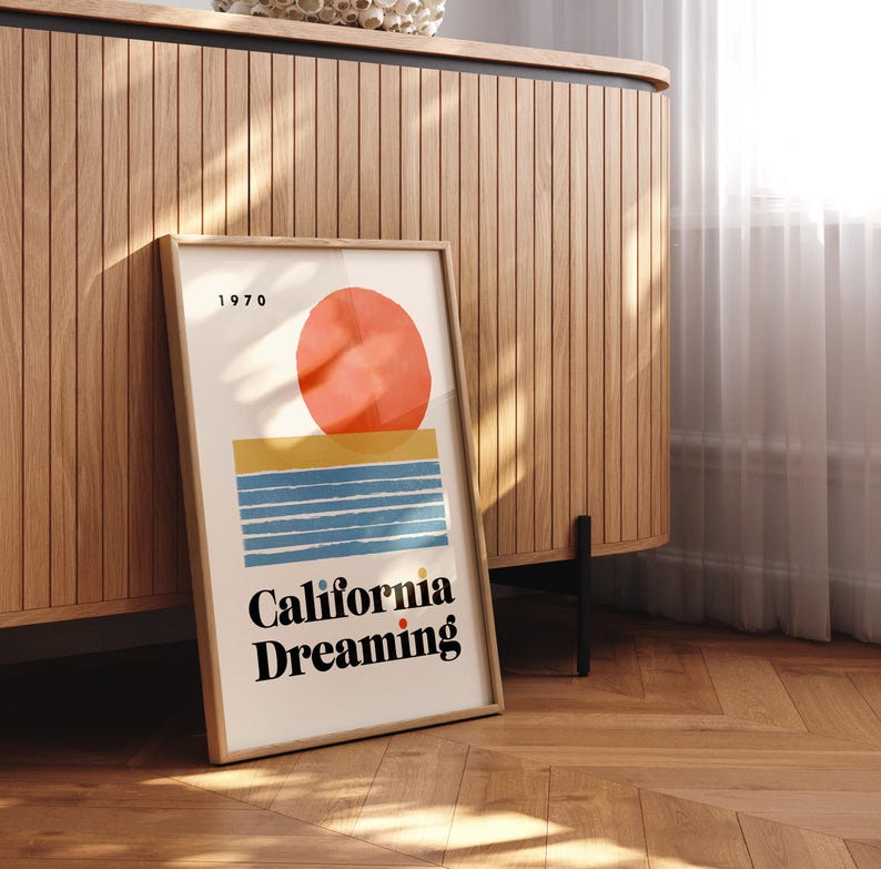 Almanac Co Prints framed poster titled 'California Dreaming' with a red sun, blue waves, and minimalist 1970s design, perfect for home decor. Framed leaning against wooden cupboard.