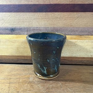 May include: A small, cylindrical ceramic cup featuring a glossy, dark brown glaze with subtle blue and green mottled accents. The vessel has a slightly flared rim and a smooth, reflective surface, showcasing a handcrafted, organic appearance.