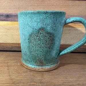 May include: A ceramic mug with a textured, speckled teal and green glaze. The mug features a prominent, curved handle and a distinct, unglazed tan base. The surface has a mottled appearance with darker brown speckles concentrated in the center.