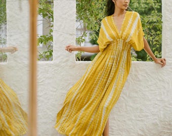 Può includere: Abito lungo giallo con motivo tie-dye bianco. L'abito ha scollo a V, maniche corte e vita arricciata. La persona è a piedi nudi vicino a un muro bianco con vegetazione verde sullo sfondo. Il tessuto è leggero.
