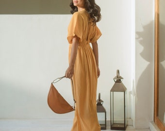 Può includere: Una donna indossa un lungo abito maxi giallo con una borsa a mano marrone a forma di mezzaluna. È in piedi in una stanza con pareti bianche e una finestra sullo sfondo.