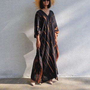 Könnte beinhalten: Eine lange, schwarze und orangefarbene Batik-Kaftan mit V-Ausschnitt. Die Kaftan wird von einer Person mit Strohhut und Sonnenbrille getragen.