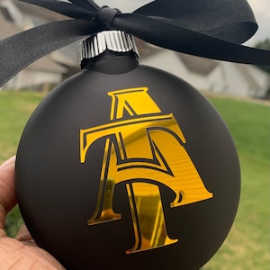 May include: Black Christmas bauble with a gold foil monogram and a black ribbon. The bauble has a matte finish and a silver metal cap. The gold monogram is a stylized "AT" design.