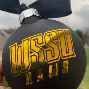 May include: A black Christmas bauble with gold lettering reading "WSSU RAMS". It has a black ribbon for hanging. The background is blurred, showing a cloudy sky and greenery.
