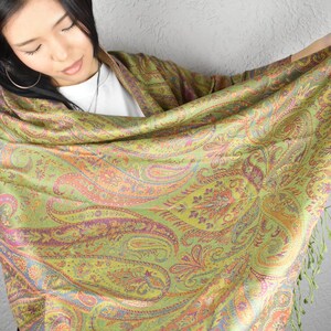 May include: A woman is holding a green and gold paisley patterned shawl. The shawl has a fringe along the bottom edge and features a detailed design with pink, orange, and purple accents. The shawl is made of a silky material.