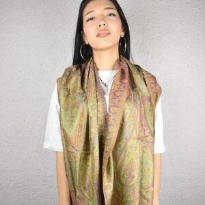 May include: A paisley patterned scarf in shades of green, orange, and purple. The scarf has a fringed edge and is draped over a white t-shirt. The scarf's design is intricate and detailed, with a classic paisley pattern.