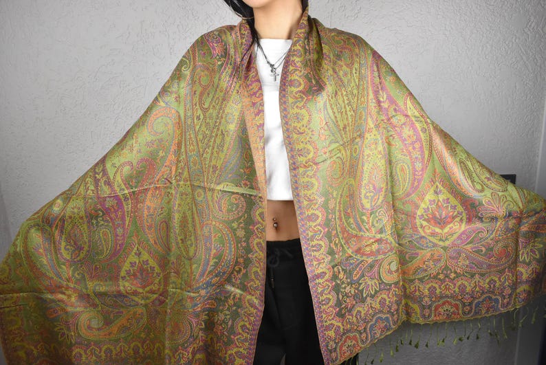 May include: A large, colorful paisley shawl in shades of green, pink, and orange. The shawl features an intricate design with a fringed edge. The fabric appears to be soft and flowing, suitable for draping.
