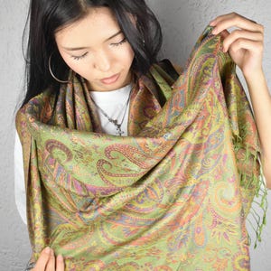 May include: A green paisley scarf with orange, purple, and blue accents. The scarf is draped and held up, showcasing the intricate design and fringe detail. The scarf is made of a soft, flowing material.