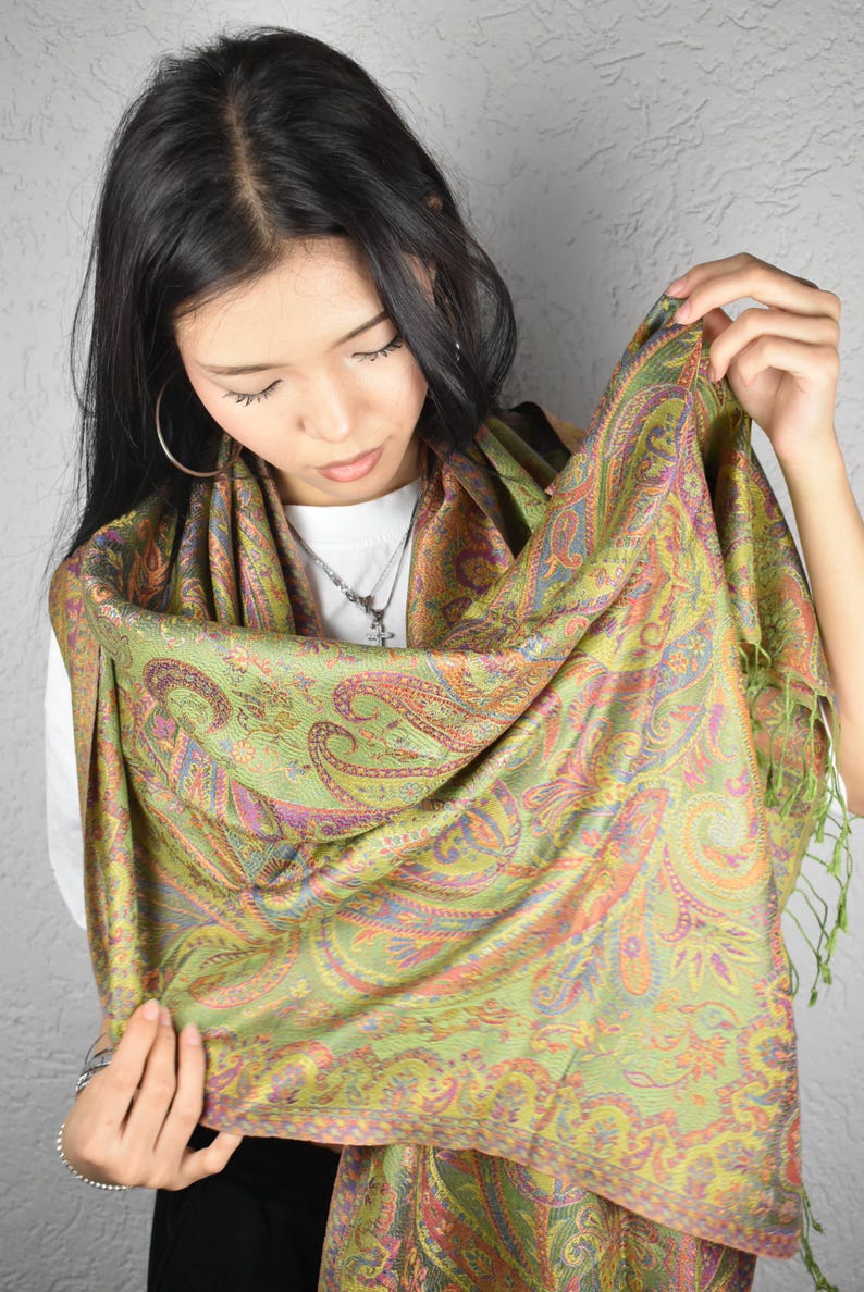 May include: A green paisley scarf with orange, purple, and blue accents. The scarf is draped and held up, showcasing the intricate design and fringe detail. The scarf is made of a soft, flowing material.