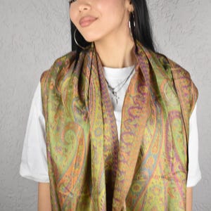 May include: A green, orange, and purple paisley patterned scarf. The scarf is draped around the neck and shoulders. The scarf is made of a soft, flowing fabric. The background is a light gray wall.