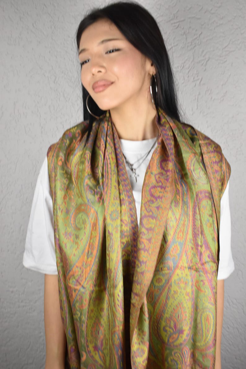 May include: A green, orange, and purple paisley patterned scarf. The scarf is draped around the neck and shoulders. The scarf is made of a soft, flowing fabric. The background is a light gray wall.