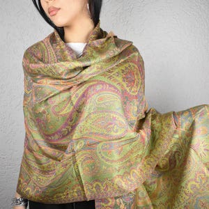 May include: A green and gold paisley patterned shawl. The shawl features a detailed design with accents of pink, orange, and blue. The fabric is draped over the shoulders, showcasing the intricate pattern and texture. The shawl has a fringed edge.