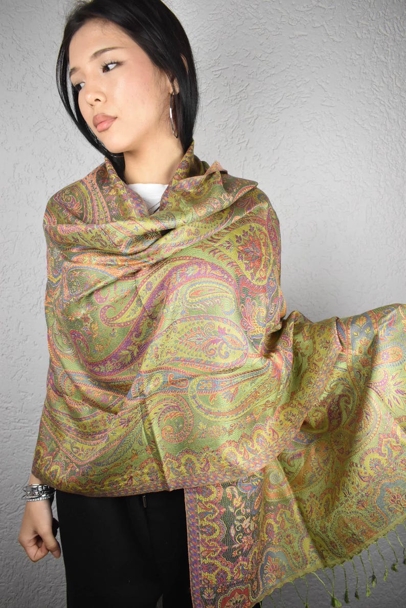 May include: A green and gold paisley patterned shawl. The shawl features a detailed design with accents of pink, orange, and blue. The fabric is draped over the shoulders, showcasing the intricate pattern and texture. The shawl has a fringed edge.