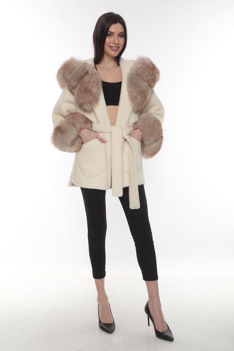 May include: A white faux fur coat with a brown faux fur trim around the hood and cuffs. The coat has a tie belt and two pockets.