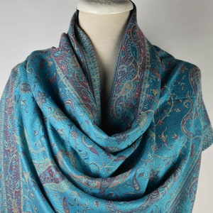 May include: A teal blue paisley patterned shawl with a maroon and gold border. The shawl is draped over a white mannequin.