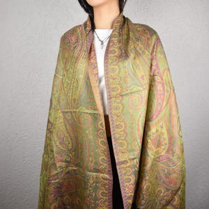 May include: A paisley patterned shawl in shades of green, pink, and orange. The shawl is draped over the shoulders, showcasing the intricate design and fringed edges. The textile appears to be soft and lightweight, suitable for layering.