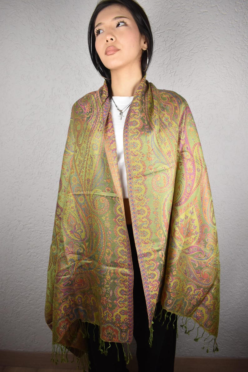 May include: A paisley patterned shawl in shades of green, pink, and orange. The shawl is draped over the shoulders, showcasing the intricate design and fringed edges. The textile appears to be soft and lightweight, suitable for layering.