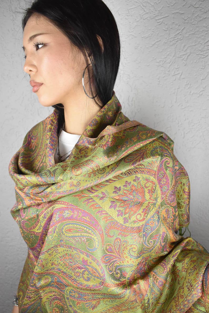 May include: A paisley patterned scarf in shades of green, orange, and purple. The scarf is draped over the shoulders, showcasing the intricate design. The fabric appears soft and flowing, suitable for a shawl or wrap.