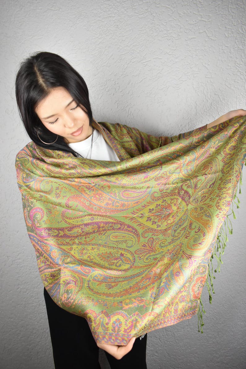 May include: A woman is holding a green and gold paisley patterned shawl. The shawl has a fringe along the bottom edge and features a detailed design with pink, orange, and purple accents. The shawl is made of a silky material.