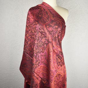 May include: A paisley patterned shawl in shades of burgundy, pink, and orange is draped over a mannequin. The intricate design features floral and curved motifs. The shawl is made of a soft, flowing fabric, perfect for adding a touch of elegance to any outfit.