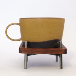 May include: A ceramic mug with a matte mustard yellow upper section and a dark brown base, featuring a thin handle. The mug rests on a square wooden platform supported by four slender metal legs that extend upward through the wood base.