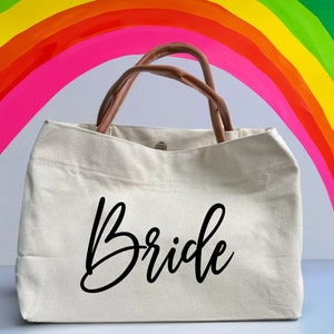 May include: A white canvas tote bag with brown leather handles. The bag has the word "Bride" written in black cursive on the front.