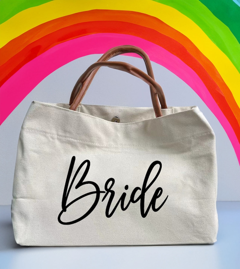 May include: A white canvas tote bag with brown leather handles. The bag has the word "Bride" written in black cursive on the front.