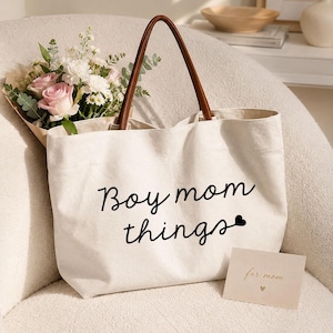 May include: A cream-colored canvas tote bag with brown leather-like handles. The front features the black printed text "Boy mom things" followed by a small black heart. A small rectangular card with the text "for mom" sits in front.